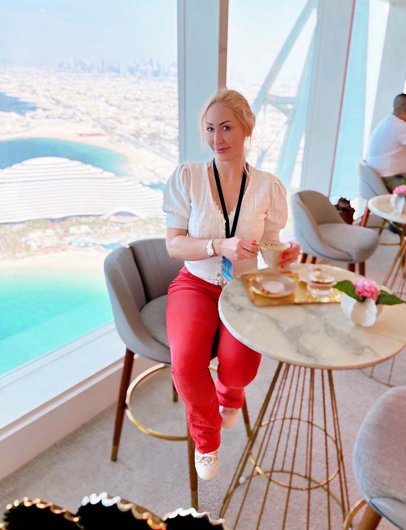 Joelle Monroe in front of a panoramic window with the Dubai skyline in the background.